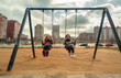 Two children playing in urban playground while wearing gas masks by toxic pollution of air. Female friends having fun sitting on swings with large buildings of contaminated town in background.