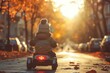 A little boy in a pompom hat and jacket rides a blue toy quad bike along an evening autumn alley with yellow trees and a sunset in the background.