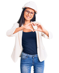 Wall Mural - Young beautiful latin girl wearing architect hardhat and glasses smiling in love doing heart symbol shape with hands. romantic concept.