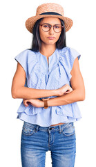 Poster - Young beautiful latin girl wearing summer hat and glasses skeptic and nervous, disapproving expression on face with crossed arms. negative person.