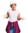 © Krakenimages.com - Young african american woman with braids wearing hardhat and builder clothes clueless and confused expression with arms and hands raised. doubt concept.