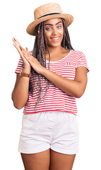 Wall Mural - Young african american woman with braids wearing summer hat clapping and applauding happy and joyful, smiling proud hands together