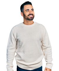 Wall Mural - Young hispanic man with beard wearing casual white sweater looking away to side with smile on face, natural expression. laughing confident.