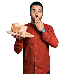 Canvas Print - Young hispanic man with beard holding bread covering mouth with hand, shocked and afraid for mistake. surprised expression