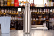 One metal vacuum-insulated bottle for water or coffee and a cup of coffee on a table on a bar background.