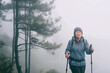 Senior Asian middle-aged woman goes hiking in the mountains, foggy morning in the mountains after rain, holding Nordic walking poles in her hands, senior active lifestyle