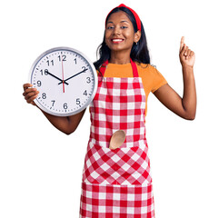 Wall Mural - Young indian girl wearing cook apron holding big clock surprised with an idea or question pointing finger with happy face, number one