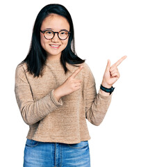 Wall Mural - Young chinese woman wearing casual sweater and glasses smiling and looking at the camera pointing with two hands and fingers to the side.