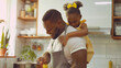 A smiling man with tattoos is cooking with a young girl in a sunlit kitchen.