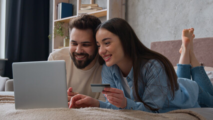 Caucasian couple happy woman and man looking at laptop screen on home bed excited smiling wife and husband pay money online use bank credit card family budget e-commerce buy goods shopping for bedroom