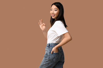 Poster - Beautiful young happy Asian woman in stylish jeans showing OK gesture on brown background