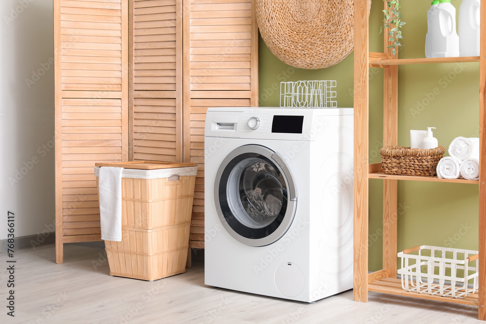 Modern washing machine with basket, shelving unit and dressing screen near green wall. Interior of home laundry room