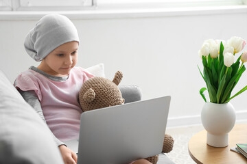 Wall Mural - Little girl after chemotherapy with toy bear using laptop at home. International Childhood Cancer Day