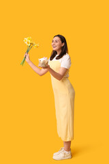 Wall Mural - Beautiful young woman with sweet Easter cake and daffodil flowers on yellow background