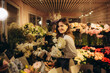 © Roman - Cheerful young florist at work. Woman picking flowers for bouquet behind a glass in a flower storage room a refrigerator. Successful owner environment friendly concept copy space