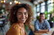 © Miss V - portrait of a curly young women sitting with colleagues behind a desk in a casual workspace, smiling
