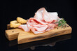 Slices of mortadella on wooden cutting board with sprig of rosemary, black pepper and salty taralli. Traditional italian starter on black reflective surface