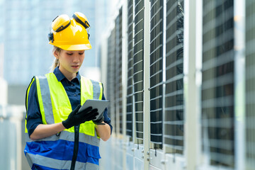 Professional Asian woman engineer in safety uniform working on digital tablet at outdoor construction site rooftop. Industrial technician worker maintenance checking building exterior air HVAC systems