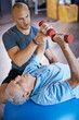 © peopleimages.com - Physiotherapist, dumbbell and old man on exercise ball for osteoporosis treatment in fitness centre, workout and pain reduction for elderly person. Male retiree, professional and senior care.