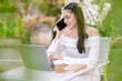 © Wasana - A young woman talking on the phone while working on a laptop in a quiet outdoor cafe.