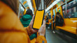 © Nataliia_Trushchenko - A woman is holding a phone in a subway car. Close-up photo of a girl's hand holding an iPhone inside a yellow subway car, with the focus on the phone's screen displaying a yellow screen