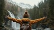 Person in yellow jacket with open arms facing a majestic waterfall in a forested mountain landscape.