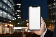 Mockup of a hand holding smart phone with blank white screen over blurred city background