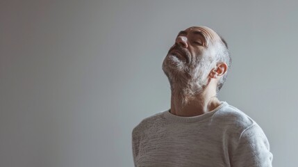 Wall Mural - Man with gray beard and hair wearing white sweater looking up with closed eyes against gray background.