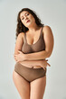 alluring plus size young girl in brown lingerie with curly hair hugging herself on grey background