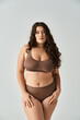 attractive plus size woman in brown lingerie with curly hair and blue eyes posing on grey background