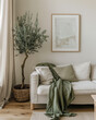 A living room with a white sofa, an olive green throw blanket on the couch and an olive tree in a pot beside it, light beige walls, a picture frame hanging above the sofa, a simple interior