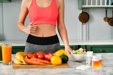 Cropped shot of slim sporty woman showing thumb up at kitchen table with organic vegetables and drinks. Fit athlete on healthy diet and eating. Calories burning, losing weight