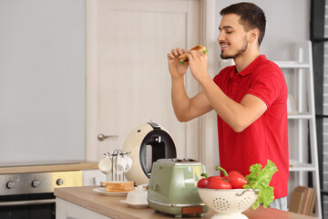 Canvas Print - Young man eating tasty sandwich made from crispy toasts in kitchen