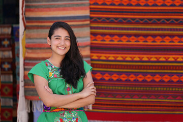 Portrait of smiling young Mexican girl dressed in typical Mexican clothing, looking at the camera with crossed arms on a background of Mexican textile handicrafts.