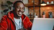A man wearing headphones and a red jacket is smiling while using a laptop