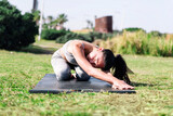 Fototapeta Las - young woman in sportswear kneeling on her yoga mat doing back stretching exercises on the grass in the park, active and healthy lifestyle concept