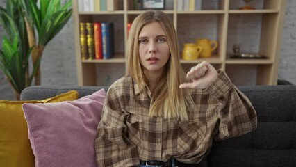 Wall Mural - Angry young blonde woman at home, showing thumbs down in negative gesture. unhappy expression while sitting on sofa, looking bad.