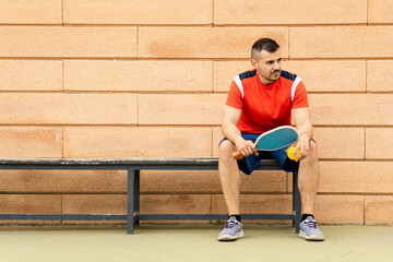 Wall Mural - An adult male in his 30s and 40s is playing pickleball on an outdoor track at sunset.The young man is sitting on a bench waiting for the game to start.Pickle ball concept.Men playing pickleball.