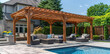 A wooden pergola with a seating area, surrounded by an outdoor pool and garden