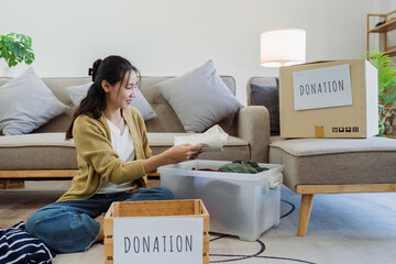 Wall Mural - Asian young woman packing clothes at home, putting on stuff into donate box with second hand clothes. Help poor people. Donation concept