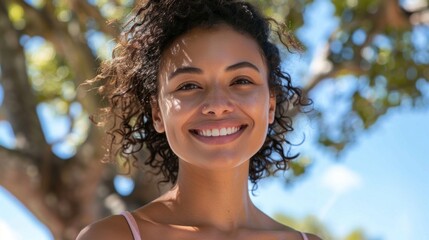 Wall Mural - A smiling woman with curly hair standing under a tree with a blue sky background.