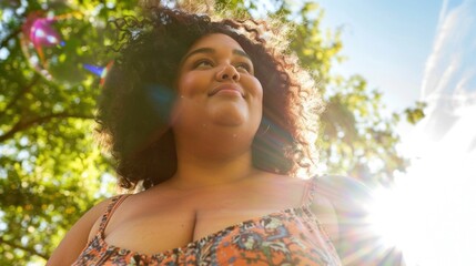Wall Mural - A joyful woman with curly hair wearing a floral top standing in a park with sunlight filtering through the leaves.