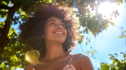 Wall Mural - A joyful woman with curly hair smiling and looking up at the sun surrounded by green leaves and a clear blue sky.