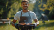 Cheerful young man holding a barbecue grill with meat in a park. Outdoor cooking and picnic concept for design and print. Summer leisure activity photography with natural background