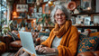 An elderly lady with grey hair and glasses is smiling gently at the camera while using her laptop. She's surrounded by lush houseplants and stylish decor in a well-lit, comfortable room.