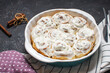 Cinnamon rolls or cinnabon, homemade recipe of sweet dessert buns with white cream sauce on dark stone background.