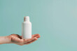 Hand presenting an empty white skincare product bottle against a pale turquoise isolated solid background, symbolizing clarity and hydration,