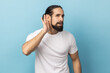 I can't hear you. Portrait of man with beard wearing white T-shirt trying to hear gossip, holding hand near ear and listening attentively carefully. Indoor studio shot isolated on blue background.