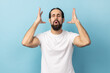 Portrait of man with beard wearing white T-shirt touching his head and showing explosion, looking worried and shocked, deadline, professional burnout. Indoor studio shot isolated on blue background.