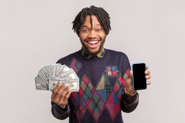 Portrait of excited happy joyful african-american man with dreadlocks and beard holding dollar banknotes and showing cell phone with empty display. Indoor studio shot isolated on gray background.